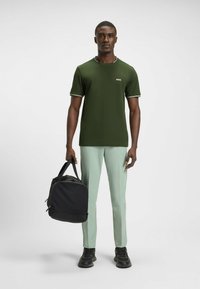 Olive green short-sleeve t-shirt with white trim, paired with mint green pants. Black duffel bag held in right hand, black sneakers worn.