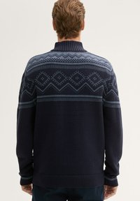 Granatowy sweter z dzianiny z wysokim kołnierzem. Posiada geometryczne wzory w jasnoniebieskim, w tym romby i zygzaki na ramionach.