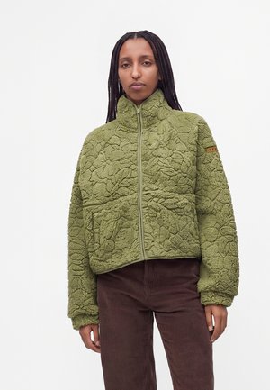 Femme aux longs cheveux tressés portant une veste polaire zippée vert olive texturée et un pantalon en velours côtelé marron foncé sur fond uni.