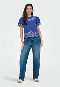 Top blu con maniche corte e decorazioni ricamate bianche lungo l'orlo e il collo, abbinato a jeans in denim dal taglio morbido e sandali leggeri.