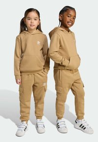 Svetlozelen hoody in trenirke, narejene iz mehkega materiala. Opremljen je s kenguru žepi in belimi logotipnimi detajli. Čevlji so beli z črnimi črtami.