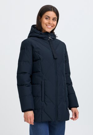 Femme souriante portant une veste d'hiver matelassée bleu marine avec capuche et fermeture éclair, debout sur un fond clair uni.