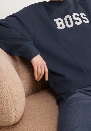 Personne portant un sweat-shirt bleu marine avec "BOSS" en lettres blanches, assise sur un canapé texturé brun clair, le bras reposant sur le coussin.