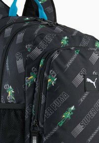 Črn nahrbtnik s zeleno in rumeno vzorčasto zasnovo, dvema zadrgama in izrazito črno nalepko logotipa Puma.