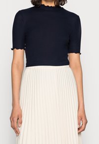Top à manches courtes côtelé bleu marine avec un col haut et des bords festonnés, complété par une jupe plissée ivoire.