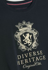 Czarna koszulka z grafiką w złotym kolorze przedstawiającą lwa, z teksturowanym wzorem oraz napisem "Różnorodne Dziedzictwo"; okrągły dekolt z czerwonymi i niebieskimi akcentami.