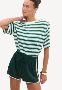 Femme portant un t-shirt rayé vert et blanc, un short vert foncé avec une bordure blanche, de grandes boucles d'oreilles dorées et un bracelet en or.