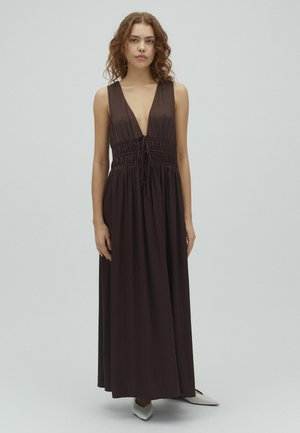 Žena s kučeravými vlasmi v tmavohnedom maxi šatách bez rukávov s hlbokým výstrihom do V a zošitým pásom, stojaca pred jednoduchým svetlým pozadím.