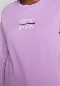 Lila sweatshirt i mjukt material med "TOMMY" i vitt och "JEANS" i röd och vit text, med horisontella randiga detaljer.