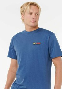 Blaue Baumwoll-T-Shirt mit kurzen Ärmeln, Rundhalsausschnitt und einem bunten "RIP CURL"-Logo auf der linken Brustseite. Glatte Textur.