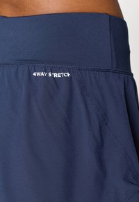 Detailní záběr na tmavě modré sportovní šortky s širokým pasem a textem "4WAY STRETCH" vytištěným blízko švu pasu.