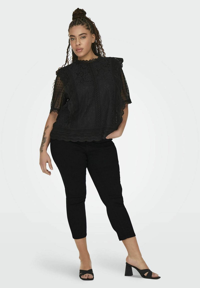Top de encaje negro con mangas abullonadas y bordes en forma de scallop, combinado con pantalones tapered negros y tacones abiertos negros. Detalles de tela texturizada visibles.