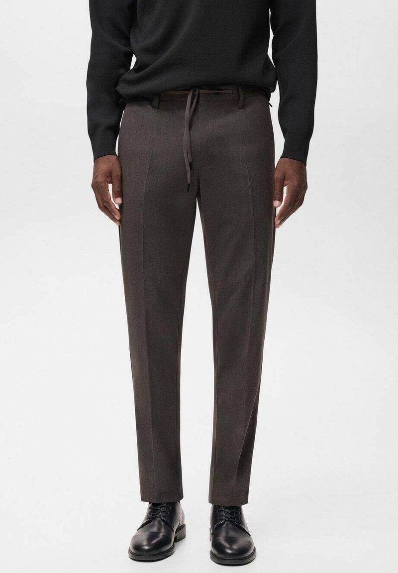 Pantaloni grigi con una texture liscia e una vestibilità slim, dotati di una vita con coulisse e sottili pieghe frontali, abbinati a scarpe nere.