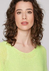 Knit sweater in bright neon green with a loose fit, round neckline, and subtle textured pattern. Curly hair frames the face.