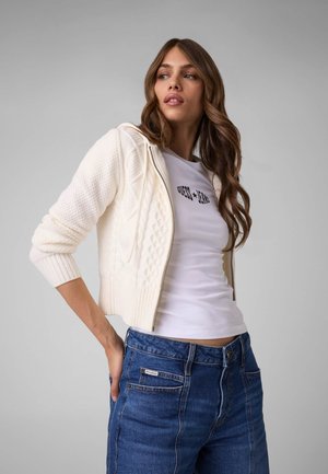 Jonge vrouw met lang golvend haar die een witte gebreide zip-up hoodie, een wit bedrukt T-shirt en blauwe spijkerbroek draagt, kijkt zijwaarts.