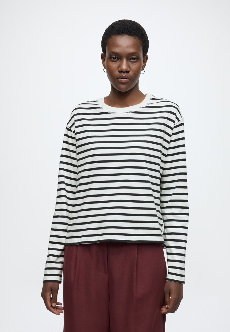 Fehér-fekete csíkos hosszú ujjú felső, laza fazonnal, kerek nyakkal és enyhén rövidített szegéllyel. Bordó nadrággal viselve.