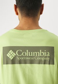 Zelené bavlněné tričko s velkým tmavě šedým obdélníkovým blokem na zadní straně, na kterém je bílé logo a ikona "Columbia Sportswear Company".