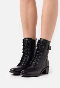 Les bottines en cuir noir présentent un design à lacets, une fermeture éclair latérale et un petit talon bloc. Les bottines ont une texture lisse et une boucle ajustable.