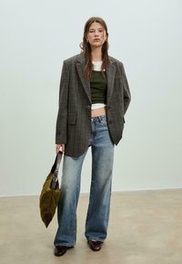 Blazer à carreaux gris sur un top vert, associé à un jean large bleu clair. Tient un sac vert texturé avec des accents foncés.