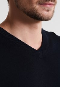 Pull col en V bleu marine foncé avec bordure en maille texturée et lignes épurées, présentant un tissu lisse et doux ainsi qu'un détail de couture subtil.