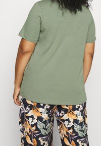 Oliivinvihreä puuvillainen T-paita, jossa on lyhyet hihat, yhdistettynä mustiin housuihin, joissa on suuret kukkakuviot oranssissa, valkoisessa ja vihreässä.