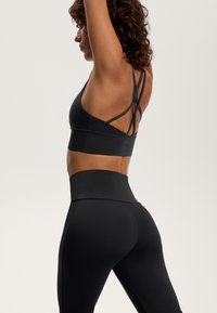 Črn športni modrček s križnimi naramnicami, v kombinaciji z visokimi črnimi legicami. Gladka tekstura, oprijet kroj in minimalno vidna blagovna znamka.