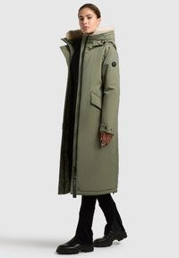 Oliven grøn lang parka med hætte foret med falsk pels, lynlåslukning, sidelommer og sorte accenter. Har en glat, polstret tekstur.