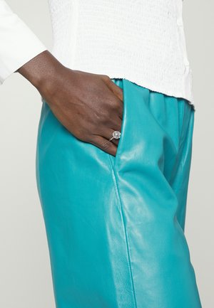 Donkere huid met een zilveren ring, rustend in de zak van turquoise leren broek, gedragen met een witte textuur top.