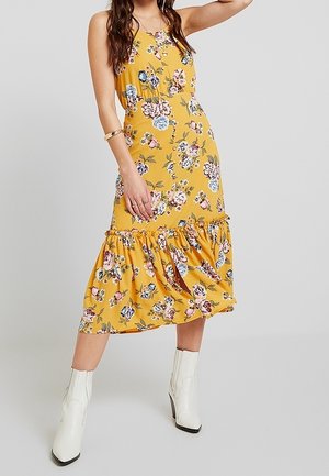 Woman in a yellow floral midi dress with ruffle hem wearing white ankle boots, posing with one arm raised against a plain background.