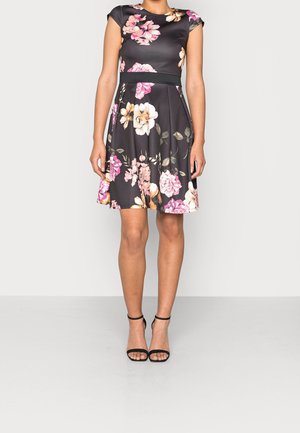 Frau trägt ein schwarzes knielanges Kleid mit rosa, weißem und beigem Blumenmuster und schwarzen Sandaletten mit Knöchelriemen, steht auf einem schlichten grauen Boden.