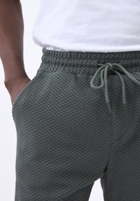 Strikkede grønne joggingbukser med elastisk talje, justerbar snor og sidelommer, med et vævet mønster overalt.