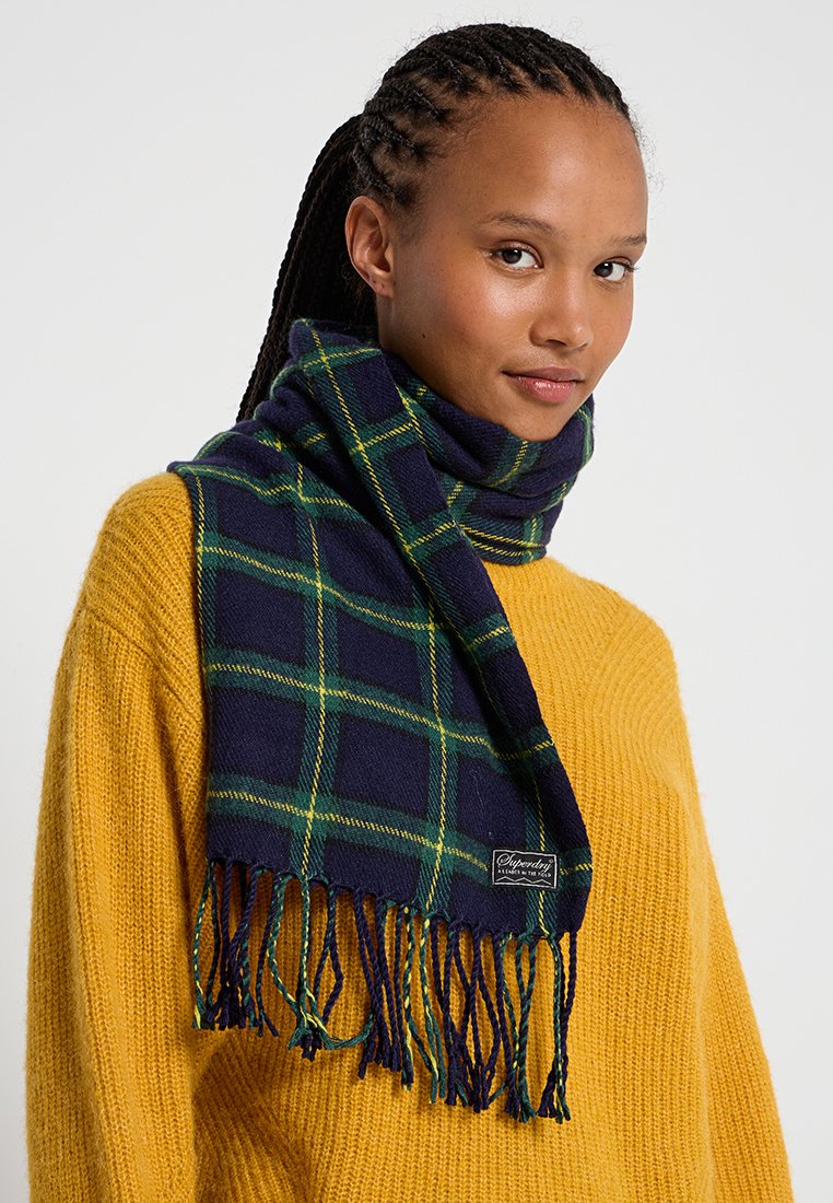 Écharpe à carreaux bleu marine avec des lignes vertes, présentant des bords frangés. Fabriquée en matériau doux, portée autour du cou, avec une étiquette de marque visible.