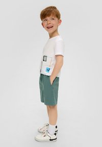 T-shirt blanche à manches courtes avec imprimés graphiques, associée à un short vert et des baskets blanches. L'ensemble est décontracté et confortable.