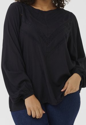 Kobieta nosząca luźną czarną bluzkę z długim rękawem z koronkowymi wzorami w kształcie litery V, do tego ciemnoniebieskie dżinsy, pozująca z ręką na biodrze.