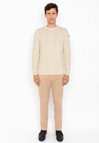 Pull beige clair à maille torsadée avec un col rond, manches longues et poignets côtelés, porté avec un pantalon beige et des chaussures noires.