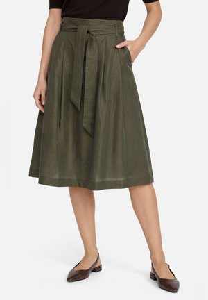Vrouw draagt een olijfgroene midi-rok met een voorstrik, zijzakken en bruine slingback schoenen met lage hak, staand tegen een witte achtergrond.
