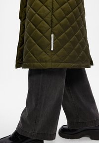 Valkots ar olīvu zaļas quilted veste ar etiķeti, kombinēta ar brīvām pelēkām biksēm un melnā platformas apavos. Vestei ir dimantu raksts un gluda tekstūra.
