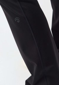 Nahaufnahme eines schwarzen Hosenbeins mit sichtbarer Naht und kleinem dunklem Logo an der Seite vor hellem Hintergrund.