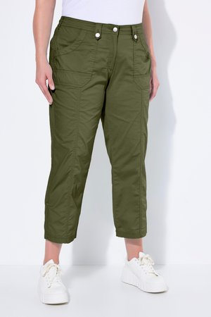 Personne portant un pantalon cargo court vert olive avec des poches avant et des baskets blanches à plateforme, debout sur un sol blanc contre un mur blanc.