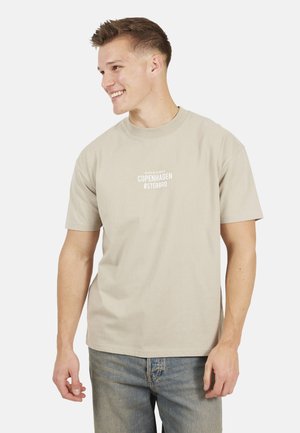 Jonge man die lacht, draagt een beige T-shirt met de tekst "Copenhagen Østerbro" en versleten blauwe jeans, staande tegen een witte achtergrond.