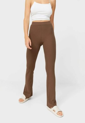 Frau trägt gerippte braune Hose mit hohem Bund, weißes bauchfreies Tanktop und weiße offene Sandalen, steht vor einfachem, hellem Hintergrund.