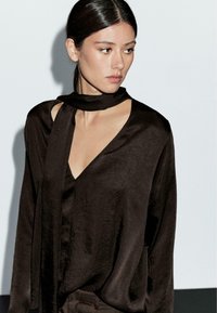 Blouse en satin brun avec un décolleté en V profond et un détail de foulard noué au col ; présente une coupe fluide et une texture subtile.
