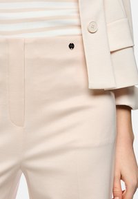 Close-up of beige tailored pants with side pocket and matching blazer sleeve over striped beige and white top.