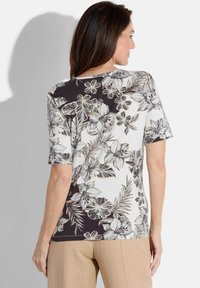 Blumenbluse mit kurzen Ärmeln, in Schwarz-Weiß-Design und einem strukturierten Stoff, der mit weißen und gold umrandeten Blumen verziert ist.