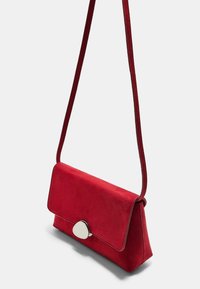 Červená semišová kabelka so štruktúrovaným tvarom, plochým prekrytím a striebornou oválnou sponou. Má dlhý, zodpovedajúci popruh na rameno.