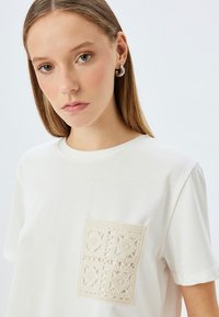 Valkoinen puuvillapaita, jossa on teksturoitu beige virkattu tasku, jossa on geometrinen kuvio vasemmalla puolella.