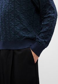 Marineblå strikket genser med geometrisk mønster og ribbestrikkede mansjetter, sammen med svarte bukser. Strukturert design over hele stoffet.