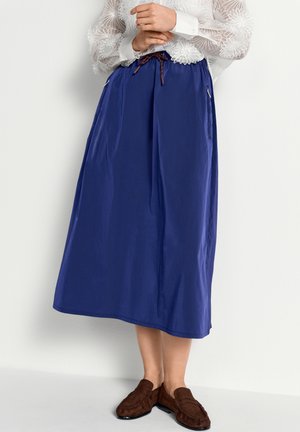 Kobieta w niebieskiej spódnicy midi z bocznymi zamkami, białej koronkowej bluzce w kwiaty i brązowych zamszowych mokasynach, stojąca z założonymi ramionami.