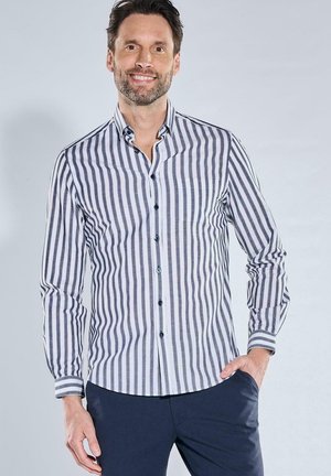 Mann lächelt, trägt ein weißes Hemd mit schwarzen vertikalen Streifen und dunkelblaue Hose, eine Hand in der Tasche, vor hellem Hintergrund.