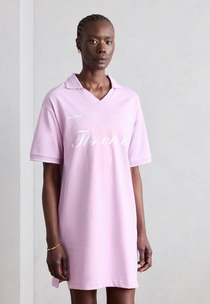 Naine palmikutega, kes kannab heledat roosat polosärki valge käsitsi kirjutatud tekstiga ja peeneid kuldseid rõngakujulisi kõrvarõngaid, seisab ühevärvilise tausta ees.